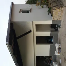 Świeżo otynkowana ściana budynku z widocznymi otworami okiennymi i drzwiami, z kilkoma wiadrami po farbie i narzędziami malarskimi w pobliżu, oraz fragmentem dachu z rynną i elementami...