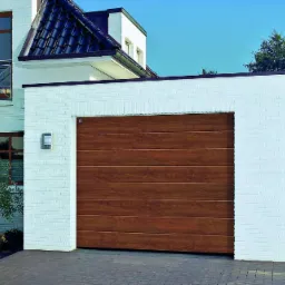 Nowoczesna brama garażowa segmentowa w kolorze drewna, zamontowana w białym, ceglanym garażu przylegającym do domu.