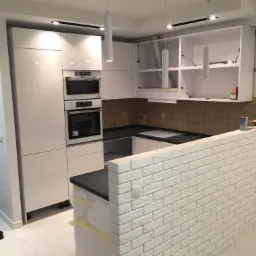 Wnętrze remontowanej kuchni z białymi, lakierowanymi frontami szafek, brązowymi płytkami na ścianie i dekoracyjną ścianką z białej cegły; widoczne wiszące lampy cylindryczne i folia ochronna...