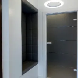 Wnęka z tapicerowanym siedziskiem i oparciem, wbudowana w białą szafę, obok szklanych drzwi z poziomymi paskami, oświetlona okrągłą lampą sufitową.