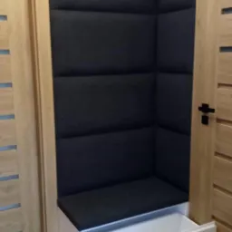 Kącik do siedzenia z tapicerowanym panelem ściennym w kolorze grafitowym i białą szafką z wysuwaną szufladą, wkomponowany między dwoje drzwi w odcieniach drewna.