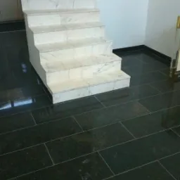 Schody wyłożone marmurem z ciemną, błyszczącą posadzką z prostokątnych płytek, widoczne odbicia światła na podłodze.