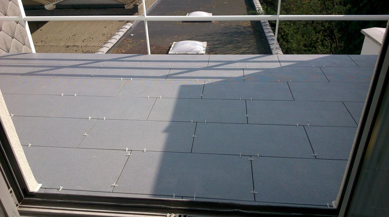 Szare płytki balkonowe imitujące drewno, ułożone z użyciem krzyżyków dystansowych, widok z góry przez otwarte okno na sąsiedni dach i zieleń.