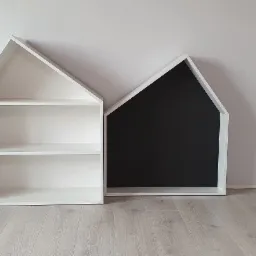 Dwa modułowe regały w kształcie domków: biały z otwartymi półkami i biały z czarnym panelem, stojące na podłodze w jasnym wnętrzu przy białej ścianie obok białych drzwi.