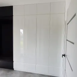 Nowoczesna szafa wnękowa z białymi, lakierowanymi frontami, połączona z czarnym segmentem z siedziskiem i wieszakami, widoczna z perspektywy wnętrza przedpokoju, z otwartymi drzwiami po prawej...