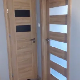 Dwa różne modele drzwi wewnętrznych z drewnianą okleiną i przeszkleniami w nowym mieszkaniu, widoczne klamki i podłoga z paneli.