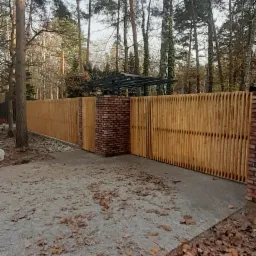 Nowoczesne ogrodzenie z pionowych drewnianych listew i ceglanych słupków na tle leśnego krajobrazu z opadłymi liśćmi.