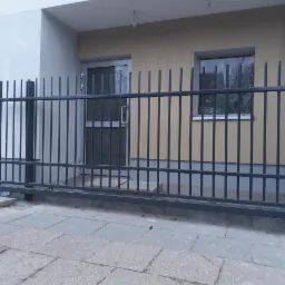 Suwana brama wjazdowa z metalowymi prętami, szare wykończenie, widoczne bloczki betonowe pod konstrukcją, szara kostka brukowa przed budynkiem.