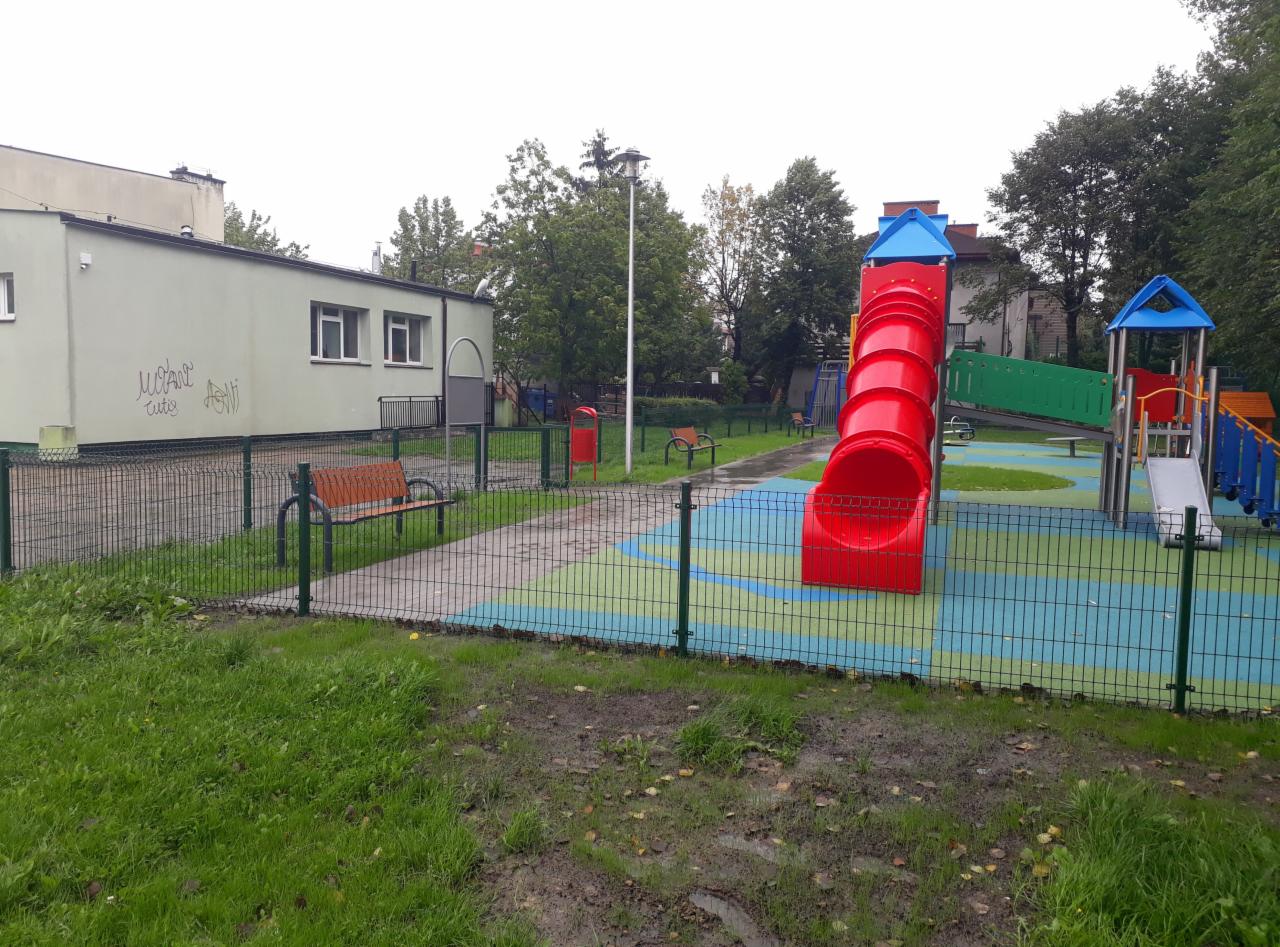 Drobnooczkowe ogrodzenie panelowe w kolorze zielonym otacza plac zabaw z czerwoną zjeżdżalnią tunelową i innymi kolorowymi konstrukcjami dla dzieci; w tle budynek z graffiti.