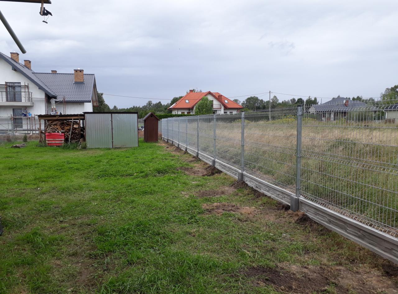 Nowo postawione ogrodzenie panelowe z podmurówką betonową, widok na dom z czerwonym dachem w tle, pochmurne niebo.