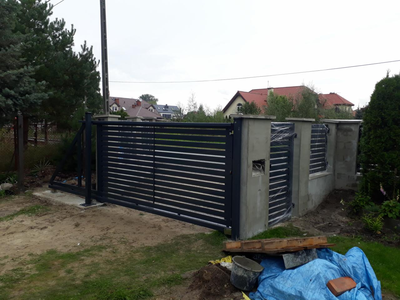 Montaż przesuwnej bramy wjazdowej z poziomymi panelami na betonowych słupkach, widoczne narzędzia i materiały budowlane, w tle domy jednorodzinne.