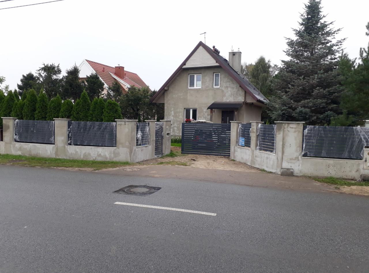 Częściowo zamontowane, grafitowe ogrodzenie z poziomych paneli, jeszcze w folii ochronnej, osadzone na betonowych słupkach, brama wjazdowa z numerem posesji 77A, w tle dom jednorodzinny.
