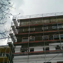 Budynek wielopiętrowy w trakcie ocieplania, widoczne rusztowania, fragment sąsiedniego budynku z balkonami i bezlistne gałęzie drzewa na tle pochmurnego nieba.