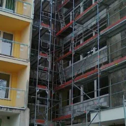 Budynek w trakcie prac elewacyjnych, widoczne rusztowania, fragmenty ocieplenia i murowane ściany między oknami, sąsiedni budynek z żółtą elewacją i balkonami.