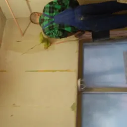 Pracownik w zielono-czarnej koszuli w kratę i niebieskich ogrodniczkach szpachluje ścianę z pęknięciem, widoczne ślady po taśmie malarskiej, okno zabezpieczone folią.