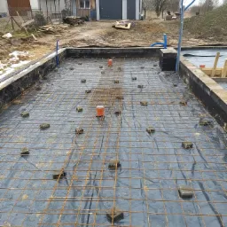 Zbrojenie płyty fundamentowej na folii hydroizolacyjnej, z dystansami z kostki brukowej, widoczne szalunki i instalacja pionowa.