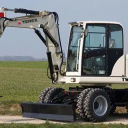 Biała koparka kołowa Terex TW70 na polu z zieloną trawą, widoczny lemiesz i ramię koparki z łyżką, opony z wyraźnym bieżnikiem.