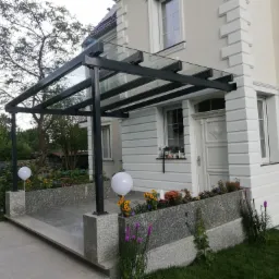 Zadaszenie nad wejściem do domu z metalową konstrukcją i szklanym wypełnieniem, oświetlone kulistymi lampami i otoczone rabatami kwiatowymi.