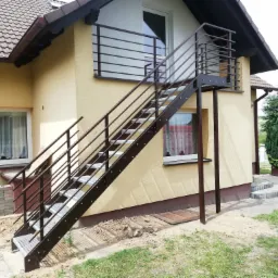 Zewnętrzne schody metalowe prowadzące do balkonu na piętrze domu, z poziomymi balustradami, widoczne cegły i ziemia przy podstawie schodów.
