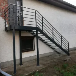 Zewnętrzne schody metalowe z ażurowymi stopniami i poziomą balustradą prowadzące do drzwi na piętrze budynku z otynkowaną elewacją.