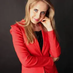 Portret uśmiechniętej blondynki w czerwonej marynarce na ciemnym tle, sugerujący profesjonalizm i pewność siebie, typowy wizerunek adwokata.