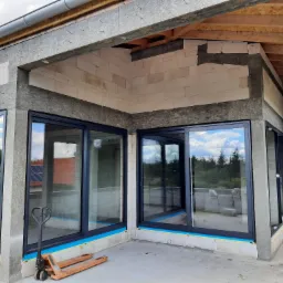 Montaż dużych, przesuwnych okien z czarnymi ramami w surowym stanie budynku, widoczny wózek paletowy i niedokończone ściany z betonu komórkowego.