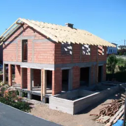 Dwukondygnacyjny dom w trakcie budowy z czerwonej cegły, z częściowo wykonanym dachem z drewnianych łat. Widoczne fundamenty i otwory okienne. Słoneczny dzień.