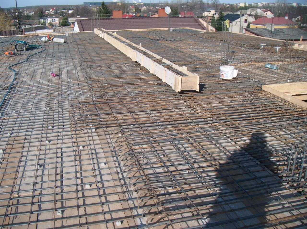 Zbrojenie stropu budynku w trakcie budowy, widoczne stalowe pręty i drewniane szalunki. W tle zabudowa miejska i błękitne niebo.