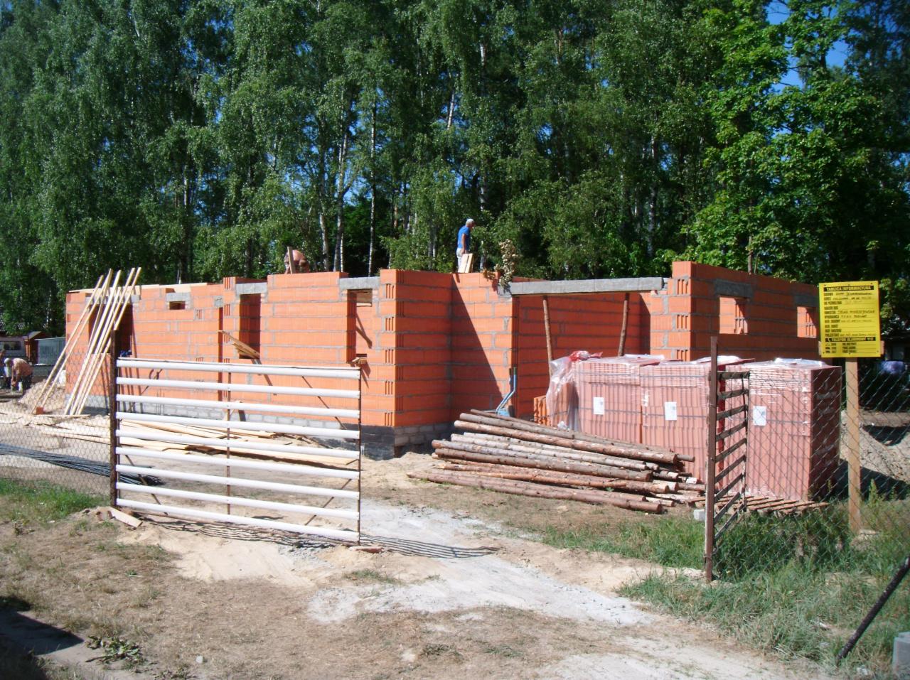 Wznoszenie ścian z czerwonej cegły na budowie domu jednorodzinnego. Widoczne palety z cegłami, drewniane podpory i ogrodzenie placu budowy. Prace murarskie w toku.