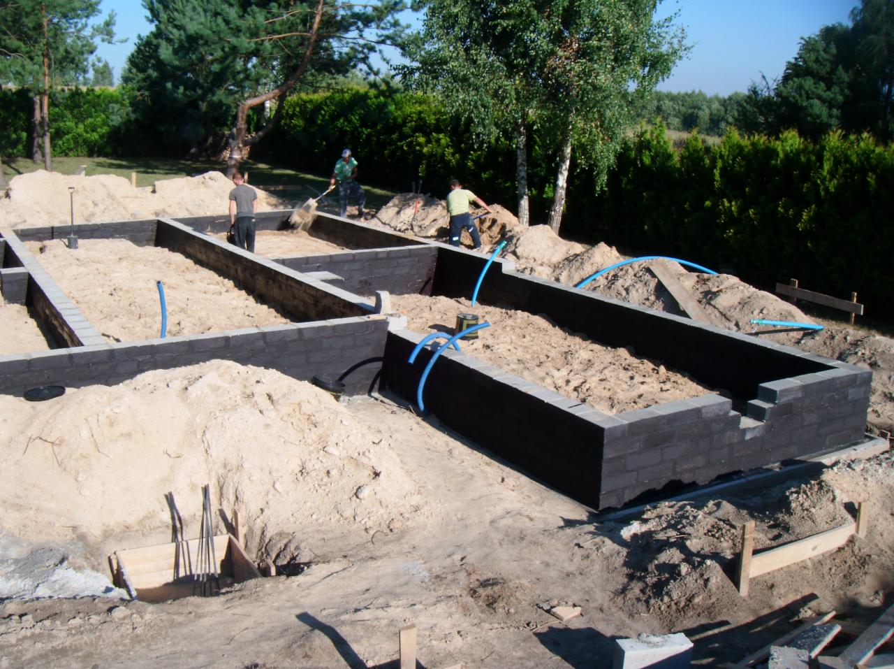 Fundamenty domu z keramzytu w trakcie budowy, widoczne ściany z bloczków i piasek wewnątrz, kilku pracowników na budowie, słoneczny dzień.