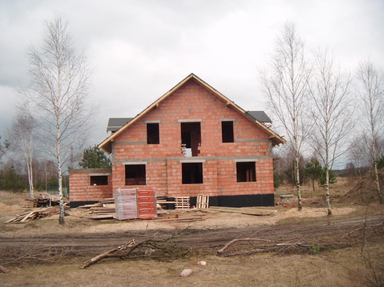 Surowy dom z czerwonej cegły w trakcie budowy, z widocznymi otworami okiennymi i stertą materiałów budowlanych, otoczony drzewami i ziemią, pochmurne niebo.