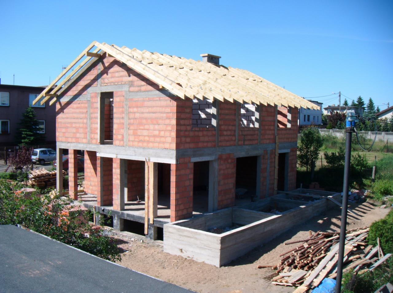 Dwukondygnacyjny dom w trakcie budowy z czerwonej cegły, z częściowo wykonanym dachem z drewnianych łat. Widoczne fundamenty i otwory okienne. Słoneczny dzień.