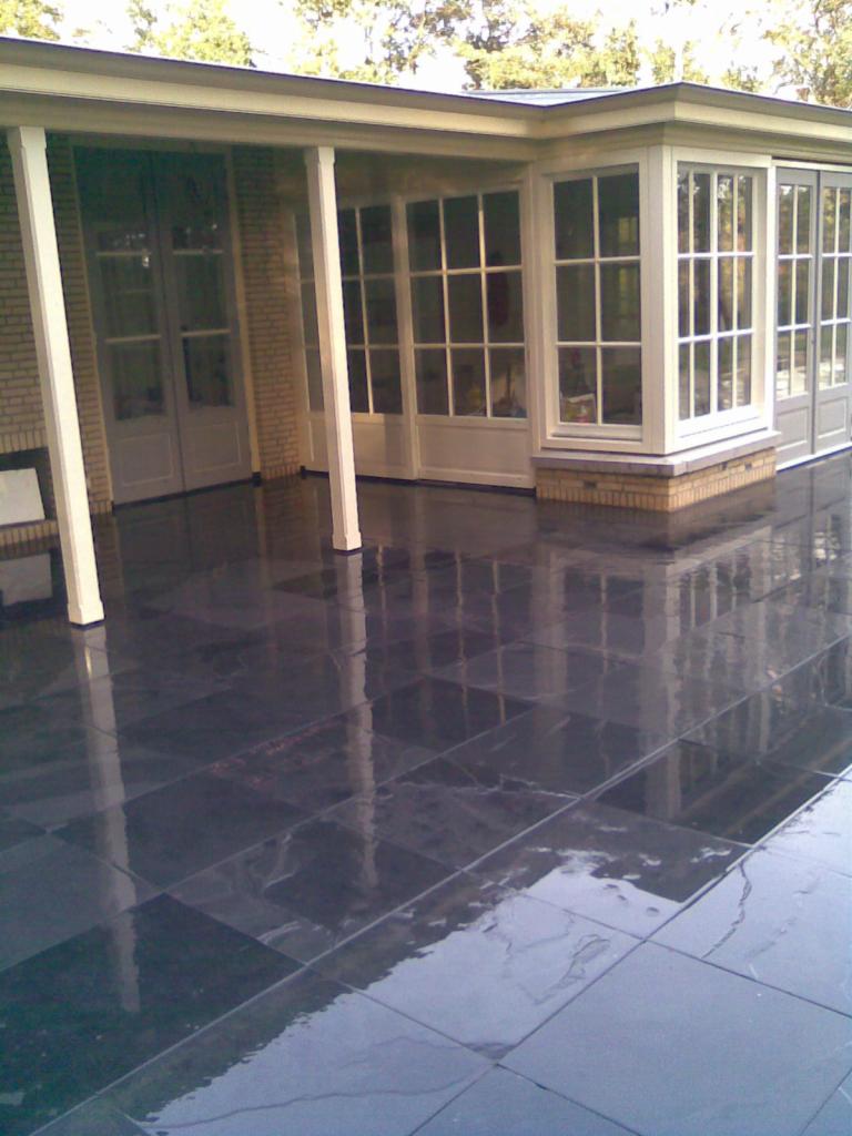 Zadaszony taras z ciemną, błyszczącą posadzką z płytek, odbijającą światło i elementy konstrukcyjne budynku.