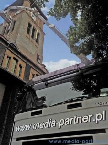 Podnośnik koszowy z wysięgnikiem sięgającym do okien starej kamienicy, widoczny fragment ciężarówki z adresem strony internetowej.