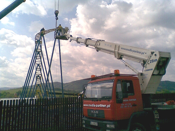 Montaż konstrukcji stalowej na wysokości przy użyciu podnośnika koszowego, widoczny krajobraz zalesionych wzgórz w tle.