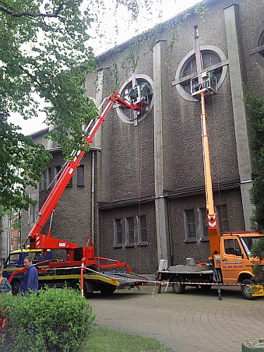 Dwa podnośniki koszowe pracują przy elewacji kościoła, montując elementy w okrągłych oknach. Jeden podnośnik jest czerwony, drugi pomarańczowy. Widoczni pracownicy w koszach podnośników.