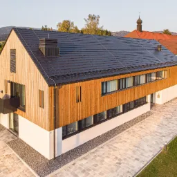 Nowoczesny budynek z elewacją z jasnego drewna i ciemną dachówką karpiówką, widok z góry na kostkę brukową w jasnych odcieniach szarości.