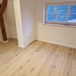 MS Innovation Bau MATEUSZ SOBCZAK - Jasna podłoga drewniana w pokoju z oknem i drewnianą belką stropową. Świeżo wykończone wnętrze z białymi ścianami i listwami przypodłogowymi.