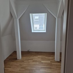 MS Innovation Bau MATEUSZ SOBCZAK - Poddasze po remoncie: drewniana podłoga, białe ściany z nietypowymi podporami i okno dachowe z widokiem na budynki.