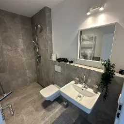 Nowoczesna łazienka z szarą, marmurkową glazurą, podwieszaną toaletą i umywalką. Widoczny prysznic, lustro i dekoracje.