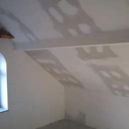 Poddasze po remoncie z oknem łukowym, widoczne ślady po malowaniu na skośnym suficie i ścianach, gniazdko elektryczne przy podłodze.