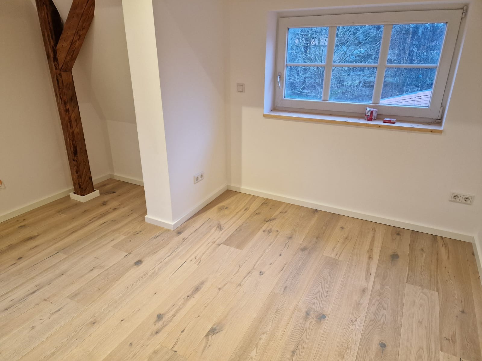 Jasna podłoga drewniana w pokoju z oknem i drewnianą belką stropową. Świeżo wykończone wnętrze z białymi ścianami i listwami przypodłogowymi.