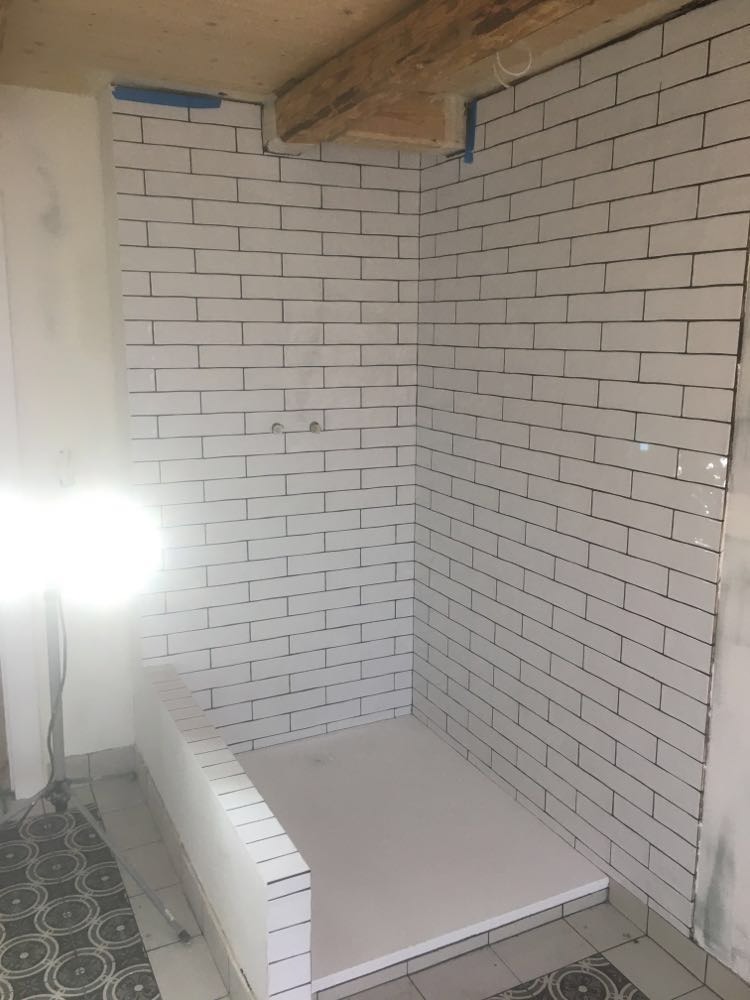 Ściany i brodzik wyłożone białymi, prostokątnymi płytkami z ciemną fugą. Widoczne drewniane elementy stropu. Fragment podłogi z wzorzystymi płytkami.