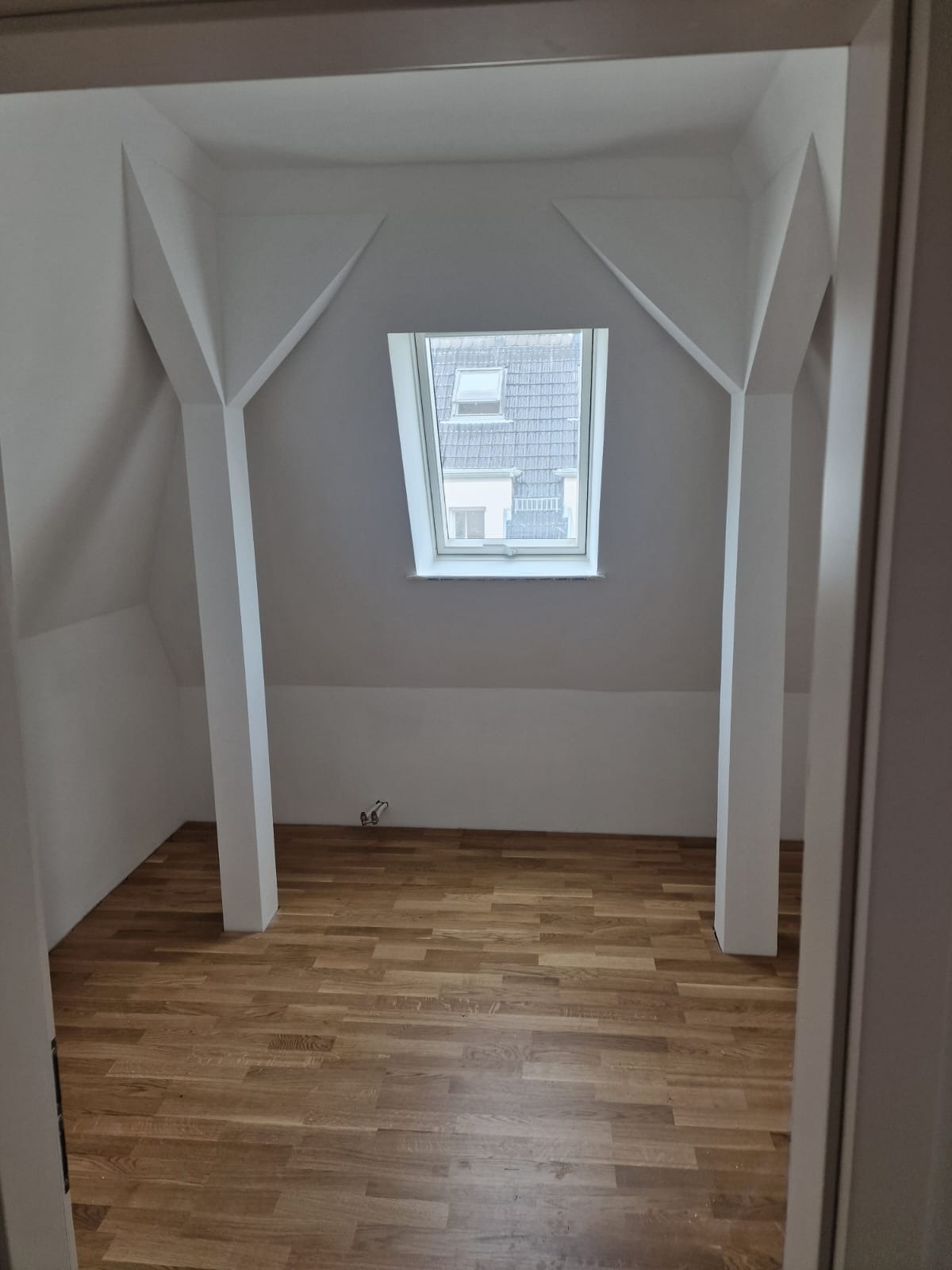 Poddasze po remoncie: drewniana podłoga, białe ściany z nietypowymi podporami i okno dachowe z widokiem na budynki.