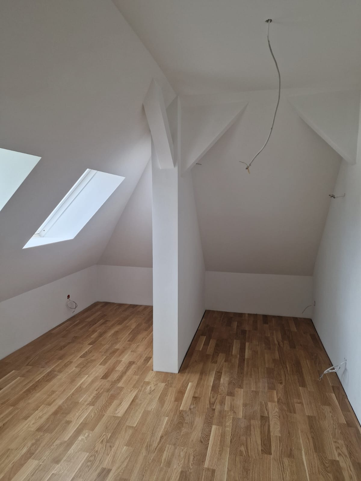 Jasne poddasze po remoncie: drewniana podłoga, białe ściany, okno dachowe i pionowa kolumna konstrukcyjna. Widoczne kable elektryczne.