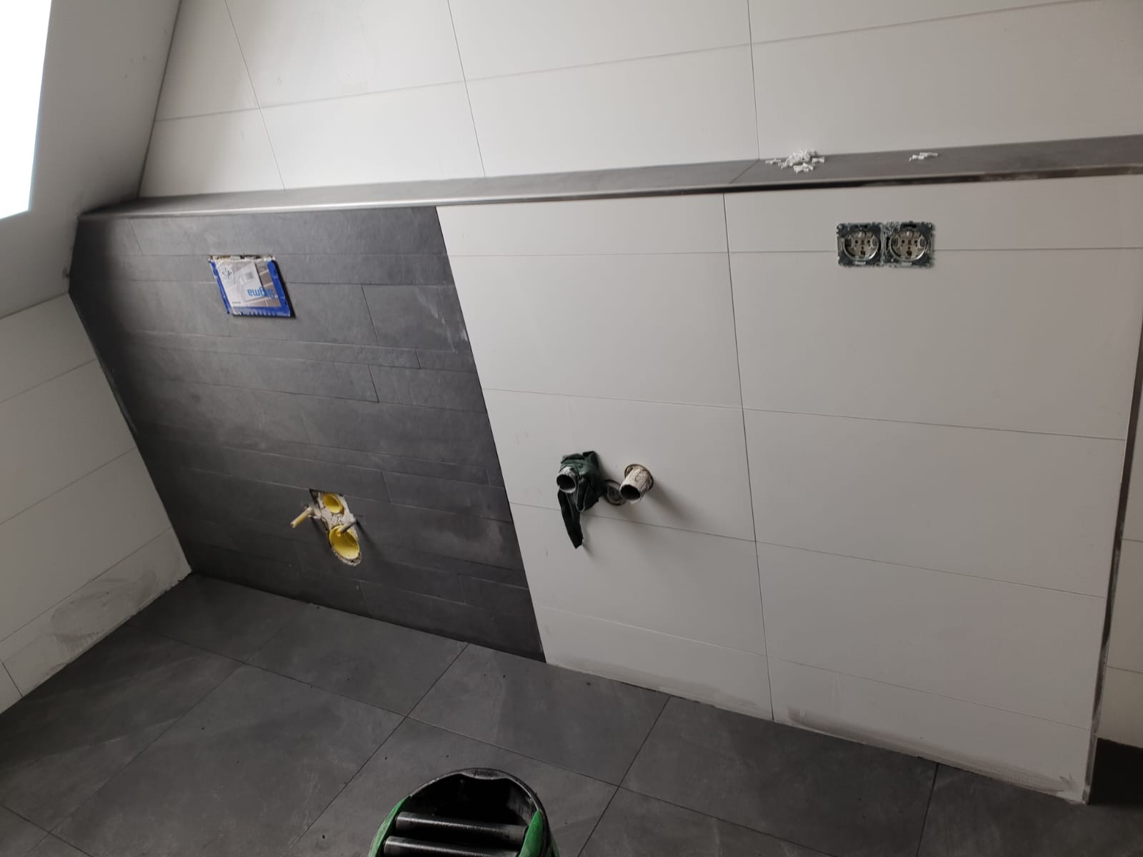 Ściana w łazience z ułożonymi płytkami w dwóch kolorach, widoczne otwory na instalacje hydrauliczne oraz elektryczne, pod skosem.