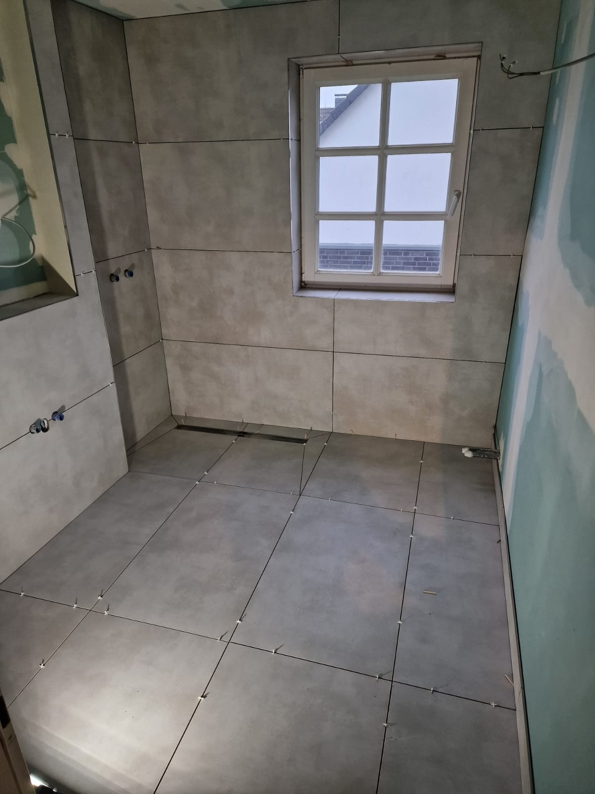 Szare płytki podłogowe i ścienne w łazience w trakcie układania. Widoczne krzyżyki dystansowe, okno i fragment instalacji. Minimalistyczny design.