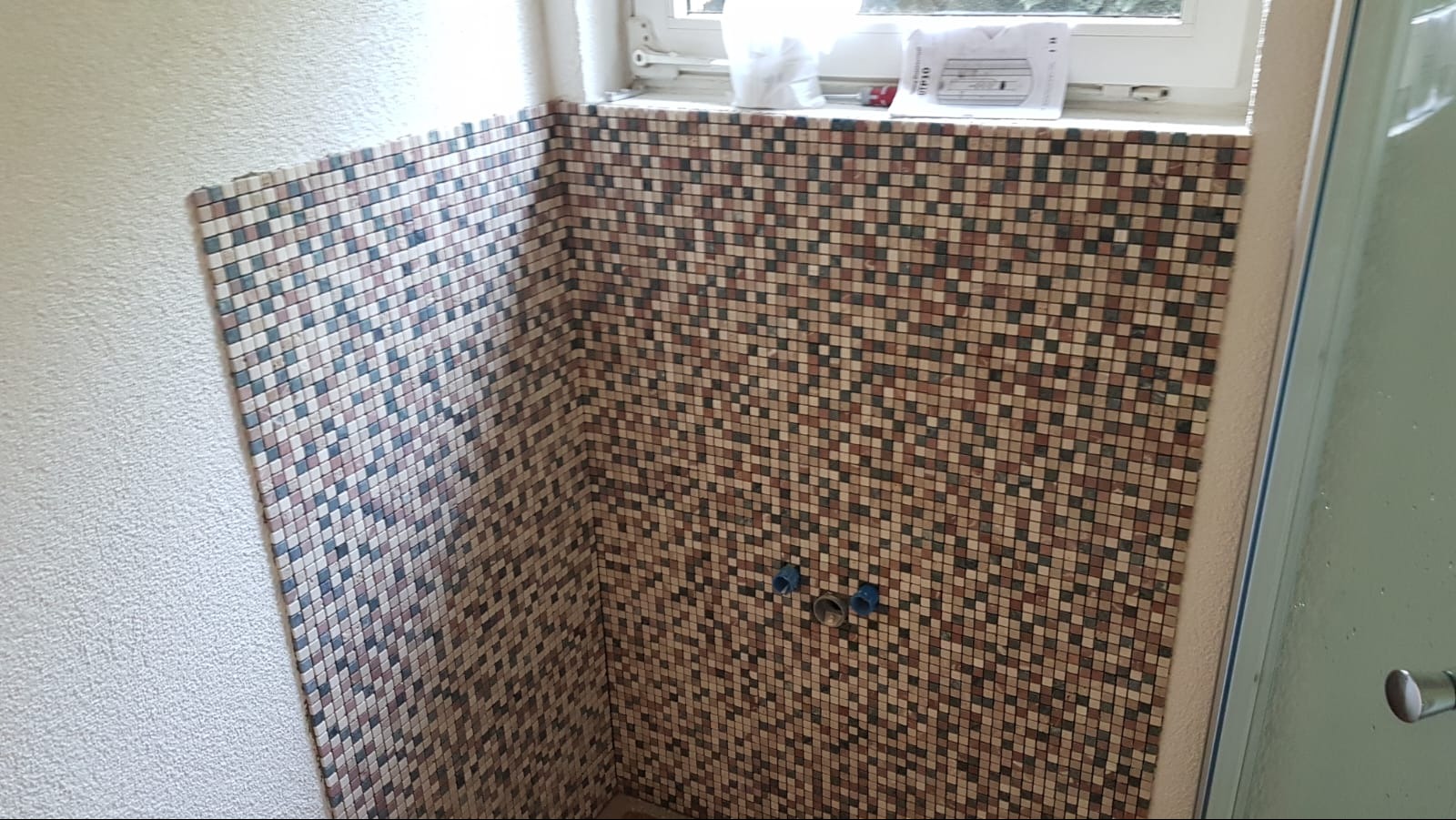 Wykończona mozaiką ściana kabiny prysznicowej z widocznymi przyłączami wodnymi i oknem w tle. Detal mozaiki w odcieniach brązu, beżu i bieli.