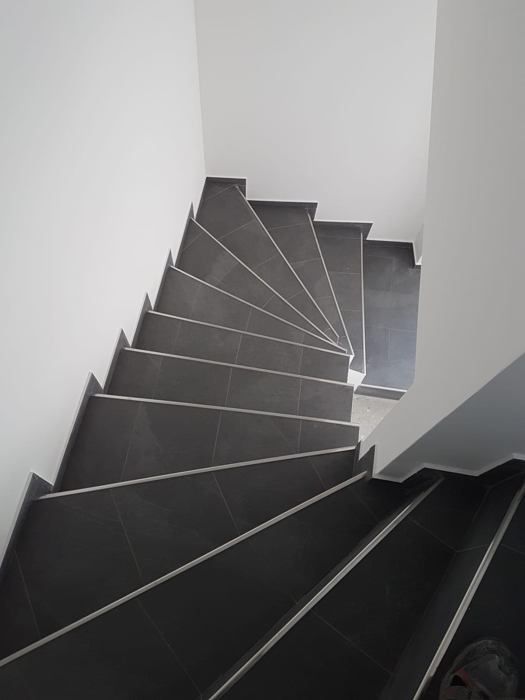 Schody wyłożone ciemną, kwadratową glazurą z jasnymi fugami, widziane z góry. Minimalistyczny design, białe ściany i precyzyjne wykończenie.