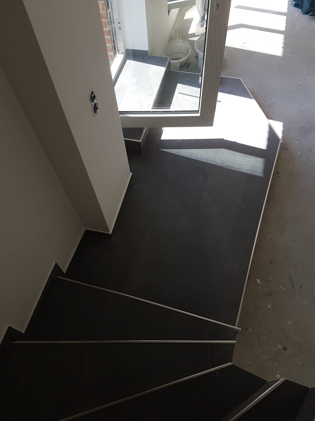 Wykończone czarną glazurą schody z metalowymi listwami antypoślizgowymi, widok z góry, fragment wejścia z widoczną puszką elektryczną w ścianie.